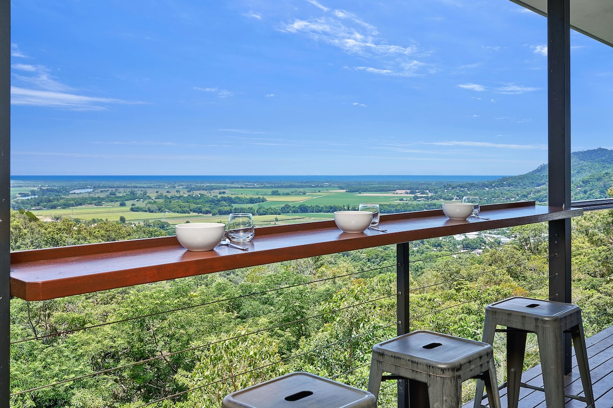 A spacious outdoor dining area is featured on a private deck, equipped with modern seating and a long wooden table. The expansive view showcases lush green landscapes and the blue coastline in the distance. Sunlight brightens the setting, creating a serene environment.