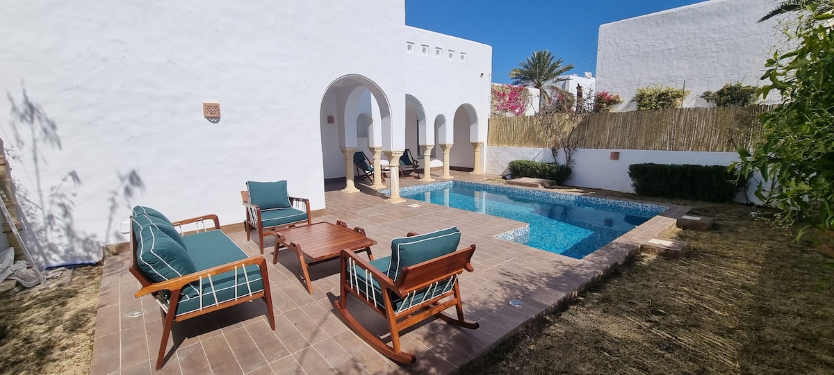 A peaceful outdoor area is presented, featuring a tranquil swimming pool surrounded by a tiled patio. Two wooden chairs and a low table are arranged nearby, complemented by lush greenery and a bamboo screen for privacy.