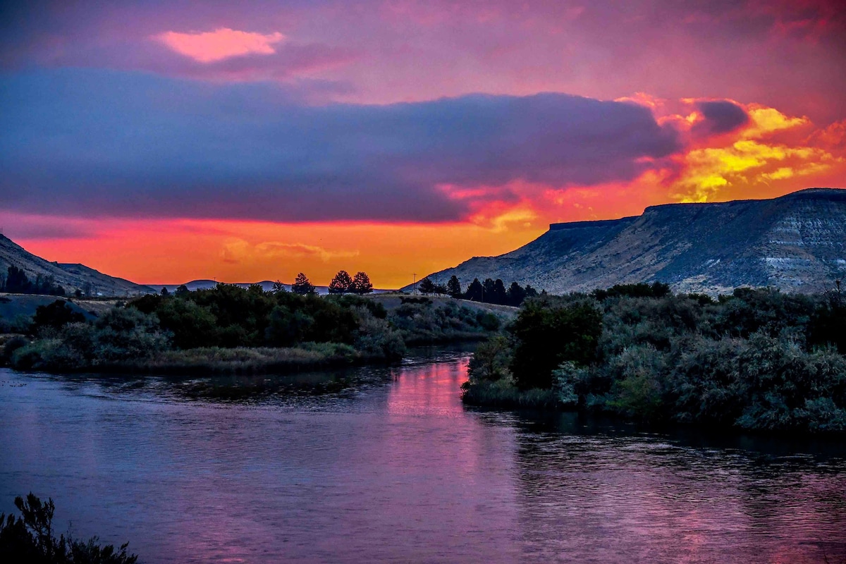 A vibrant sunset casts a colorful glow over the Snake River, reflecting shades of pink and orange on the water’s surface. Silhouetted hills frame the landscape, while lush greenery lines the riverbanks, adding depth to the serene scene.