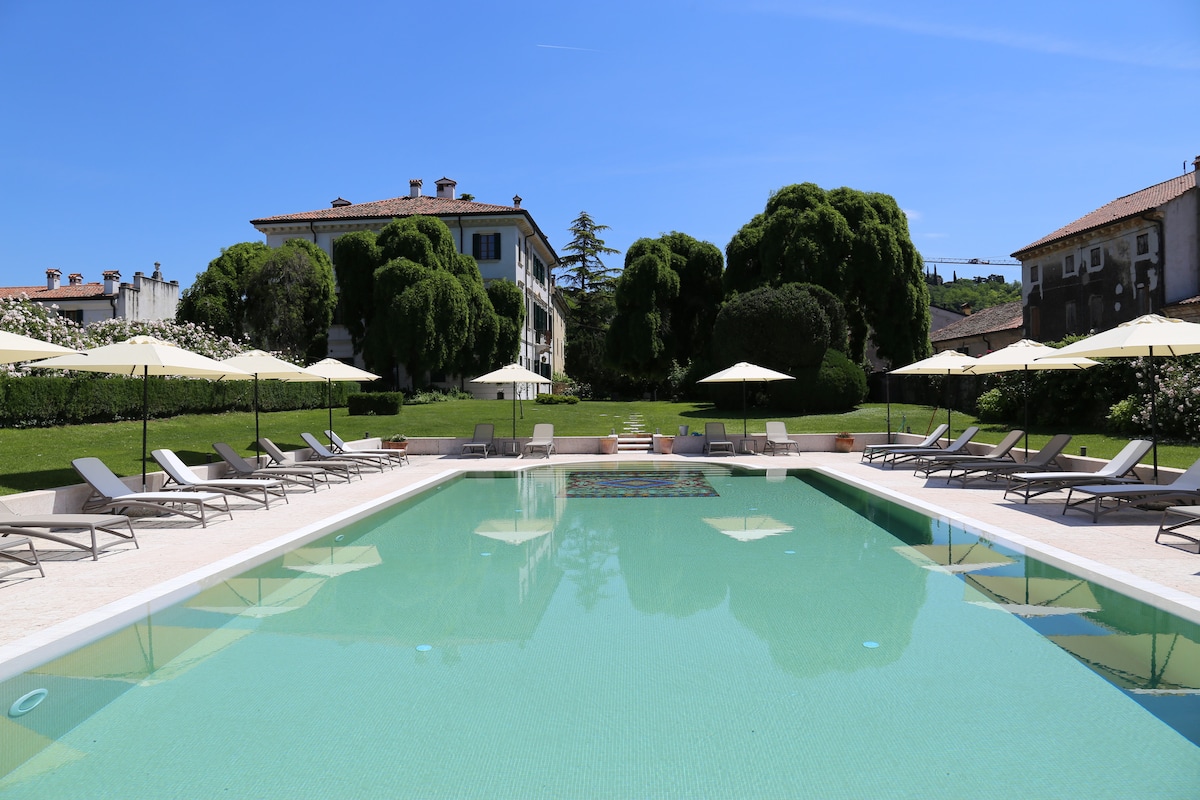 A serene outdoor swimming pool is framed by expansive green lawns and rows of lounge chairs with umbrellas. The clear water reflects the bright blue sky and surrounding greenery. Elegant villa buildings are visible in the background, contributing to a tranquil atmosphere.