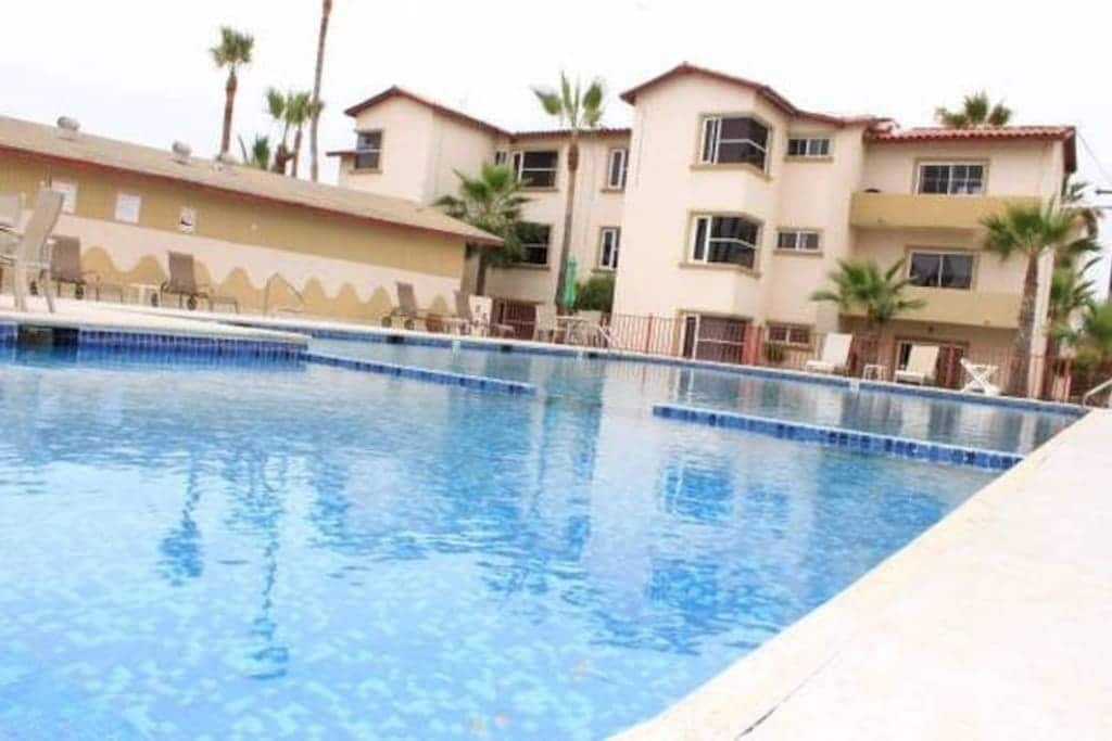 The image features a clear swimming pool, reflecting the surrounding buildings and palm trees. Multiple lounge chairs are positioned along the poolside, offering a place to relax. The backdrop consists of a multi-story condominium with a modern architecture style.