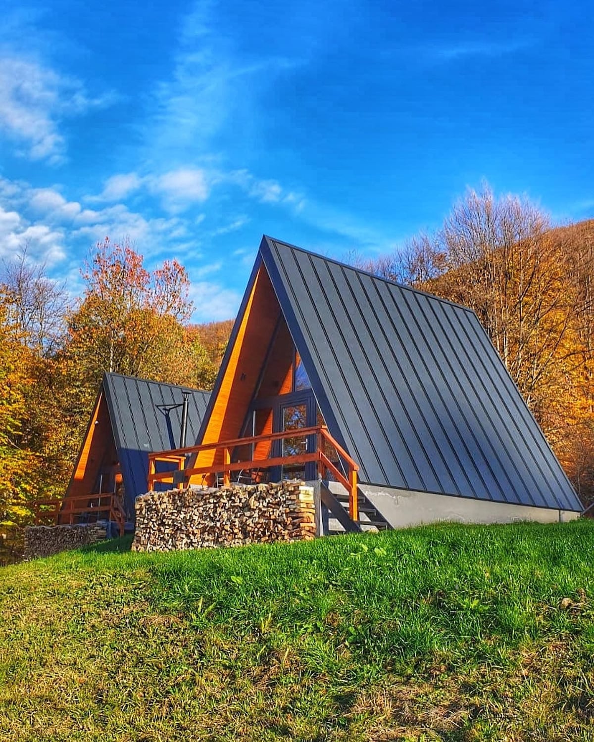 Two A-Frame cabins are positioned on a grassy slope, surrounded by autumn foliage. The structures feature steep, angular roofs with dark metal siding. A woodpile is neatly stacked beneath the deck, and a clear blue sky complements the natural setting.