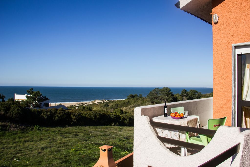 A balcony is visible, showcasing a small table set with fruit and a bottle. The coastline meets the ocean in the background, where a sandy beach stretches along the shore under a clear blue sky. Lush greenery surrounds the area.