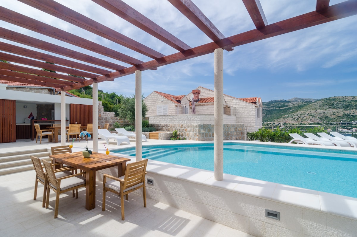 An outdoor terrace showcases a dining area with wooden furniture, positioned near a sparkling infinity swimming pool. Sun loungers surround the pool, offering a space for relaxation. The area is bordered by stone walls and lush greenery, providing a peaceful environment with open views.