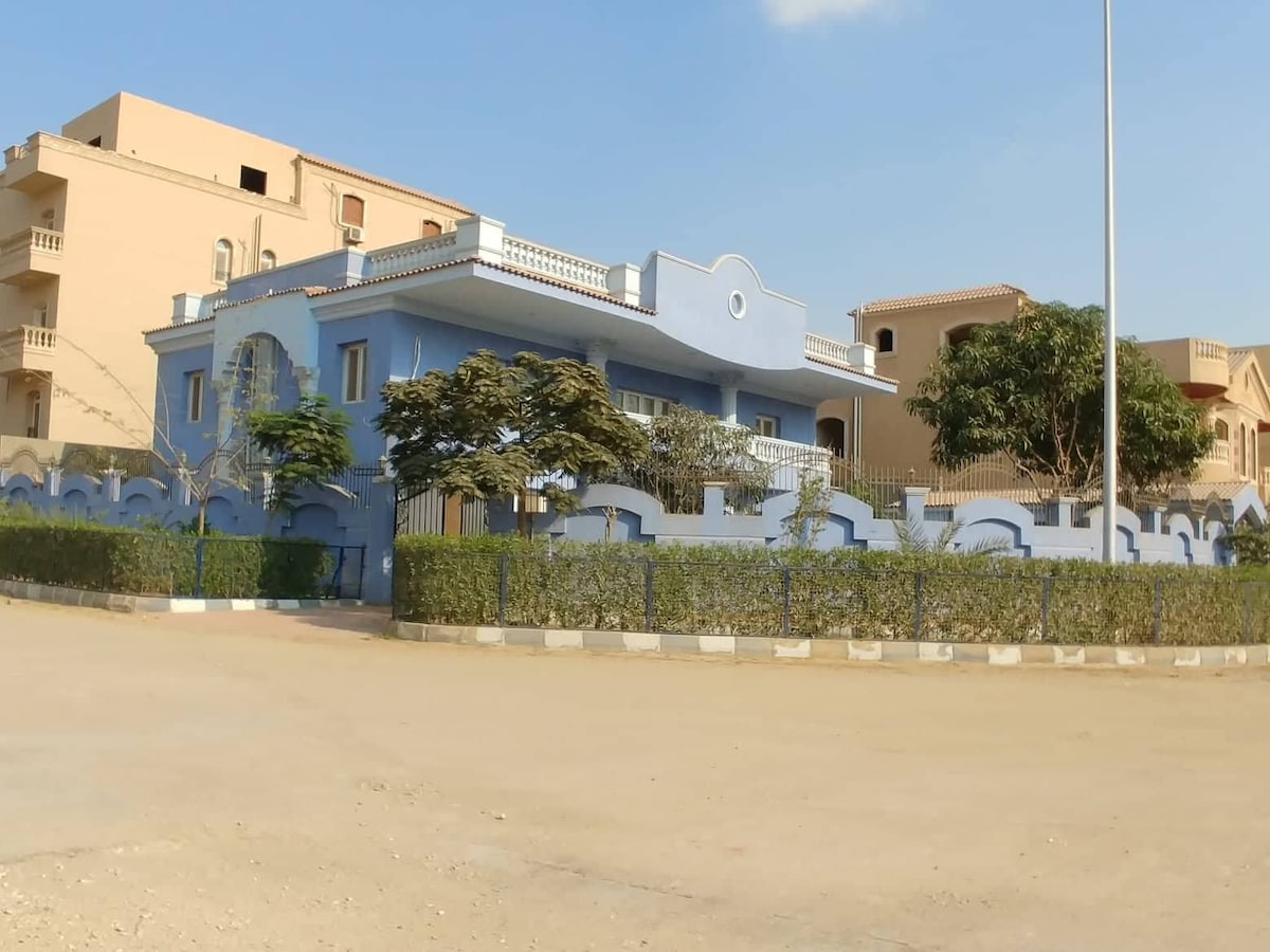 The blue villa is surrounded by neatly trimmed hedges and features a spacious front yard. Architectural details include a rounded entryway and a balcony overlooking the area. Nearby, additional residential buildings are visible under a clear sky.