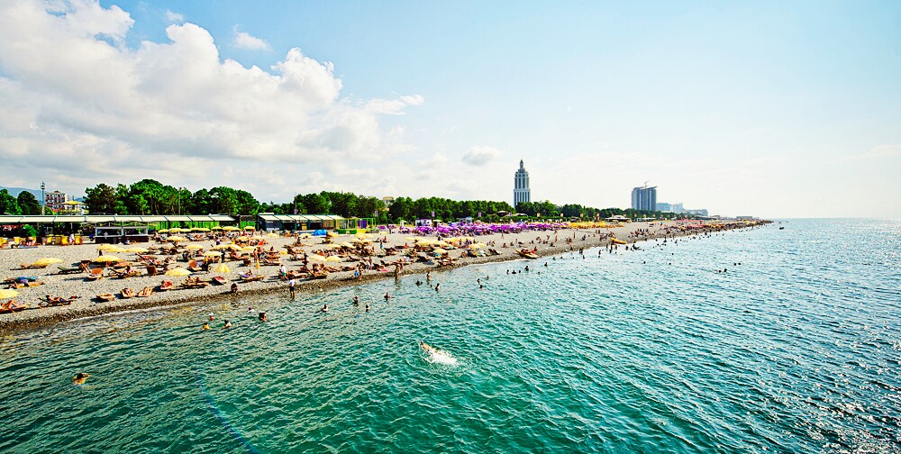 A sandy beach scene stretches along the shoreline, dotted with sunbathers and umbrellas. Gentle waves lap against the shore, while the backdrop features a mix of lush trees and urban buildings. Bright skies with scattered clouds create a lively atmosphere.