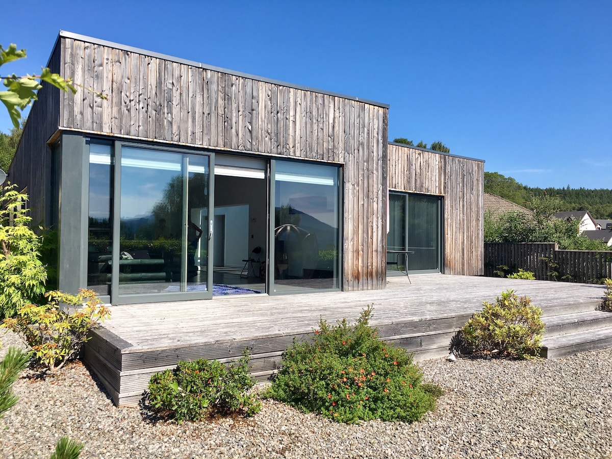 A contemporary eco house features a striking wooden facade and large sliding glass doors that open onto a spacious external decking area. The surrounding landscape includes well-maintained shrubbery and gravel pathways, creating an inviting outdoor space.