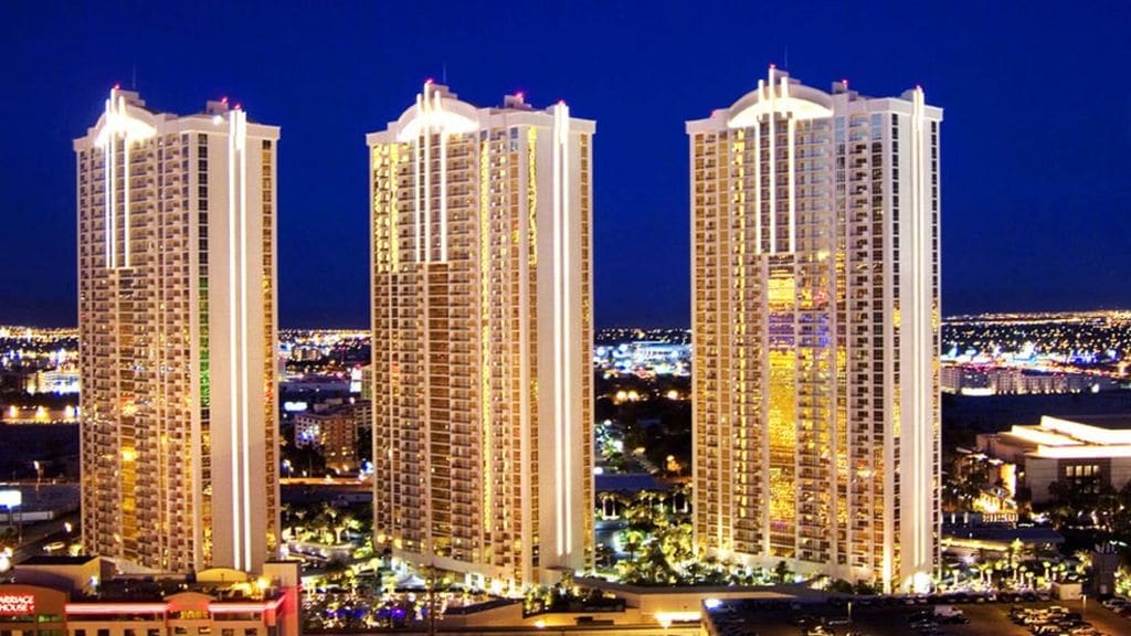 Three tall towers are illuminated against a night sky, showcasing a blend of modern architecture and vibrant city lights below. The complex is surrounded by lush greenery and offers a glimpse of the bustling Las Vegas Strip in the distance.