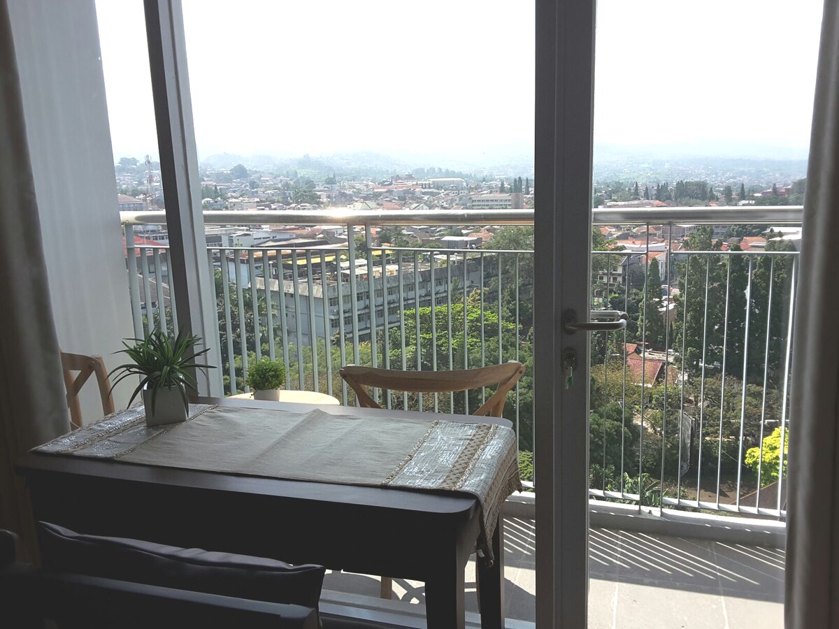 A balcony is visible through glass doors, featuring a small dining table and chairs. A view of the mountainous landscape and cityscape is captured in the background, with greenery enhancing the scenery. Natural light brightens the space, complementing the modern design.