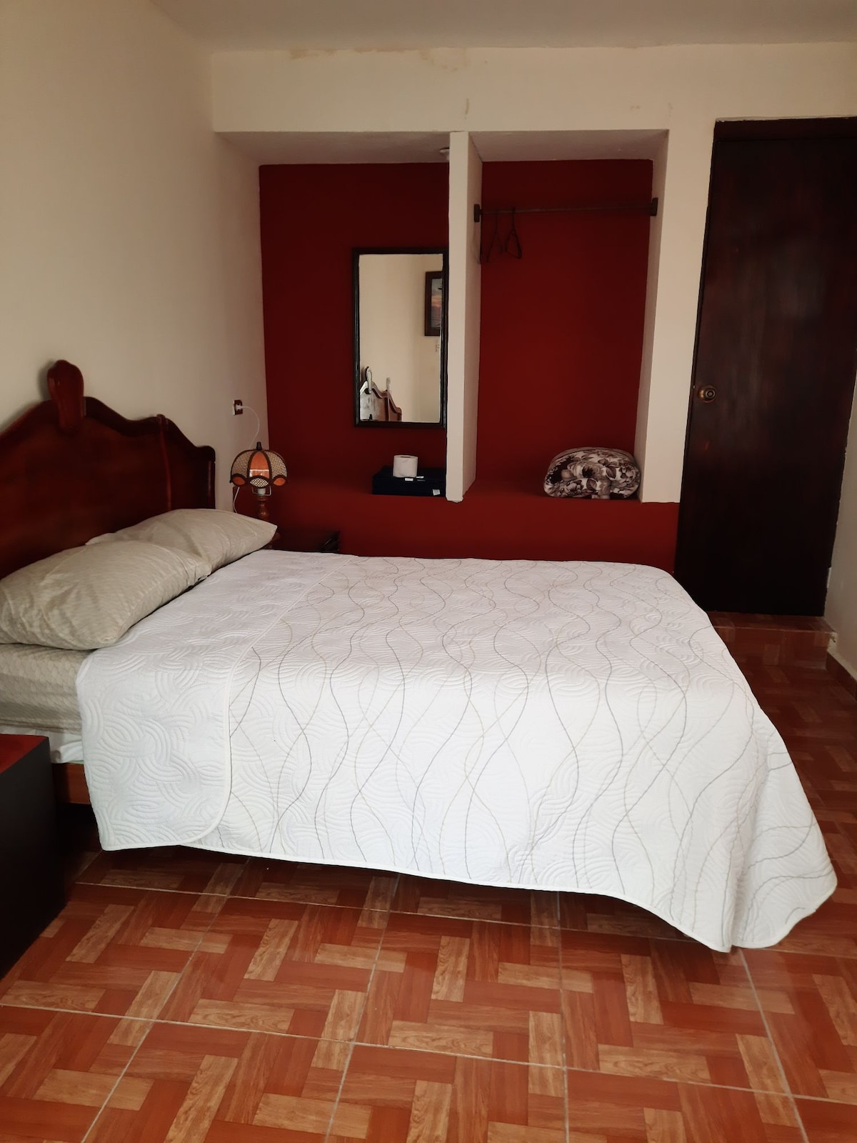 A cozy room features a spacious bed covered with a light quilt. A wooden headboard complements the warm red wall. Natural light enters through a nearby window, and a mirror and hangers are visible on the wall, providing practical storage.