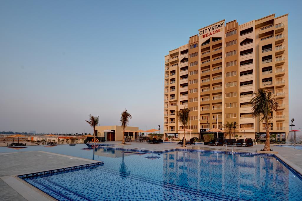 The exterior of a modern apartment building is shown, featuring a large swimming pool with tiled patterns reflecting the sky. Palm trees are situated around the pool area, alongside outdoor seating. The building is named 'CityStay Beach' across the top.