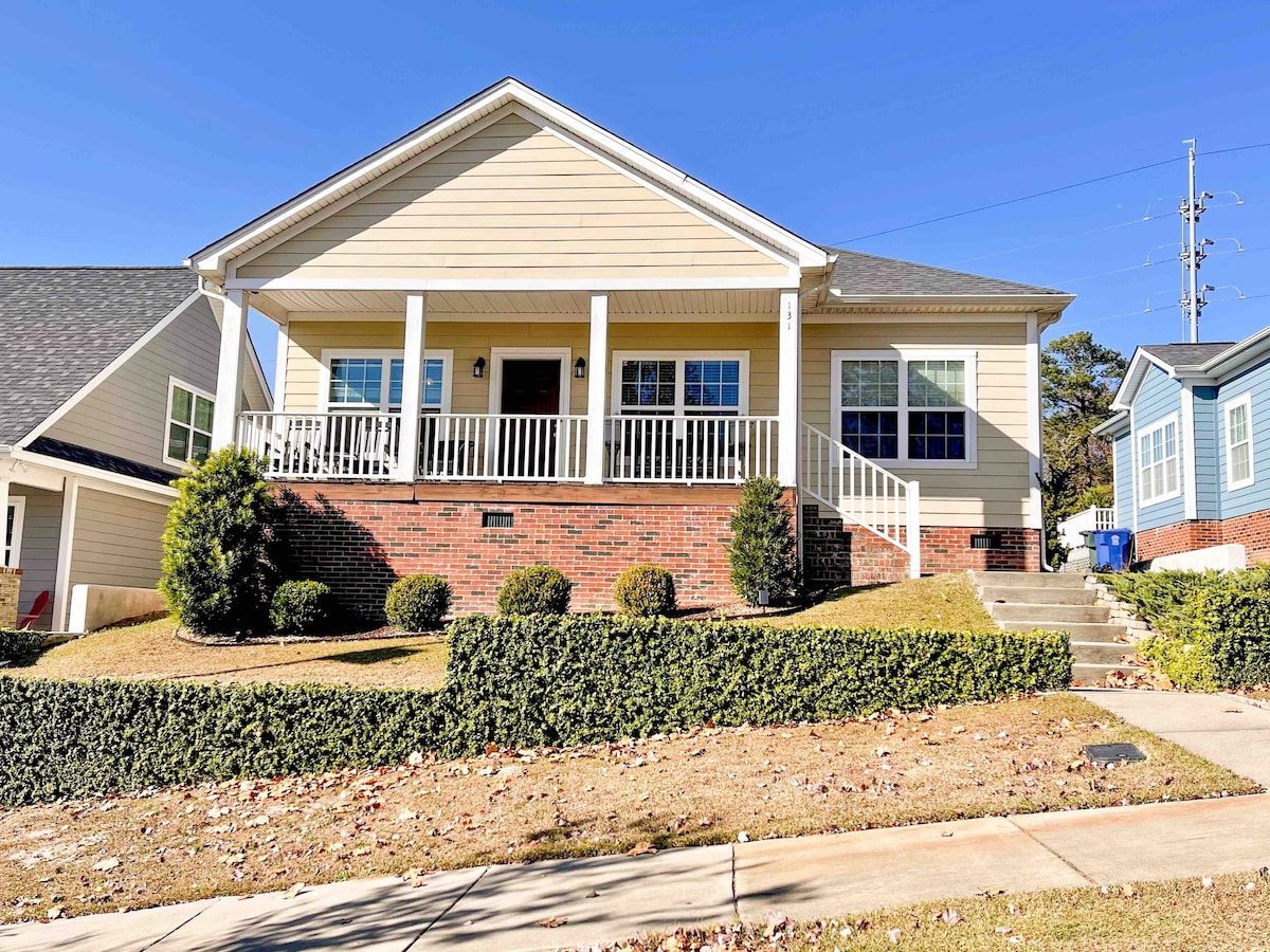 A charming bungalow is viewed from the street, featuring a wide front porch with white railings. A landscaped yard is visible, adorned with neatly trimmed bushes. The building's light-colored siding contrasts with the red brick foundation. A clear blue sky completes the welcoming scene.