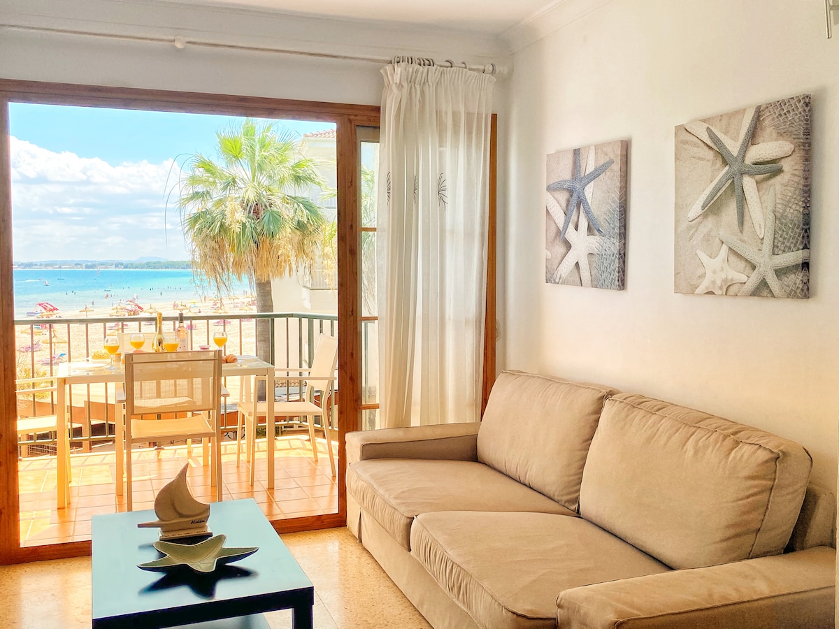 A living area is shown with a light-toned sofa facing large glass doors that open to a view of the beach and sea. Two starfish-themed art pieces are displayed on the wall, and a small table with decorative items sits in front of the sofa.