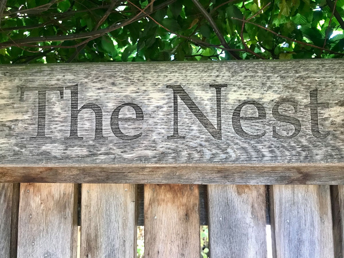 A weathered wooden sign displays the text 'The Nest,' positioned against a backdrop of lush green foliage. The natural textures of the wood are highlighted, providing a rustic charm to the entrance.