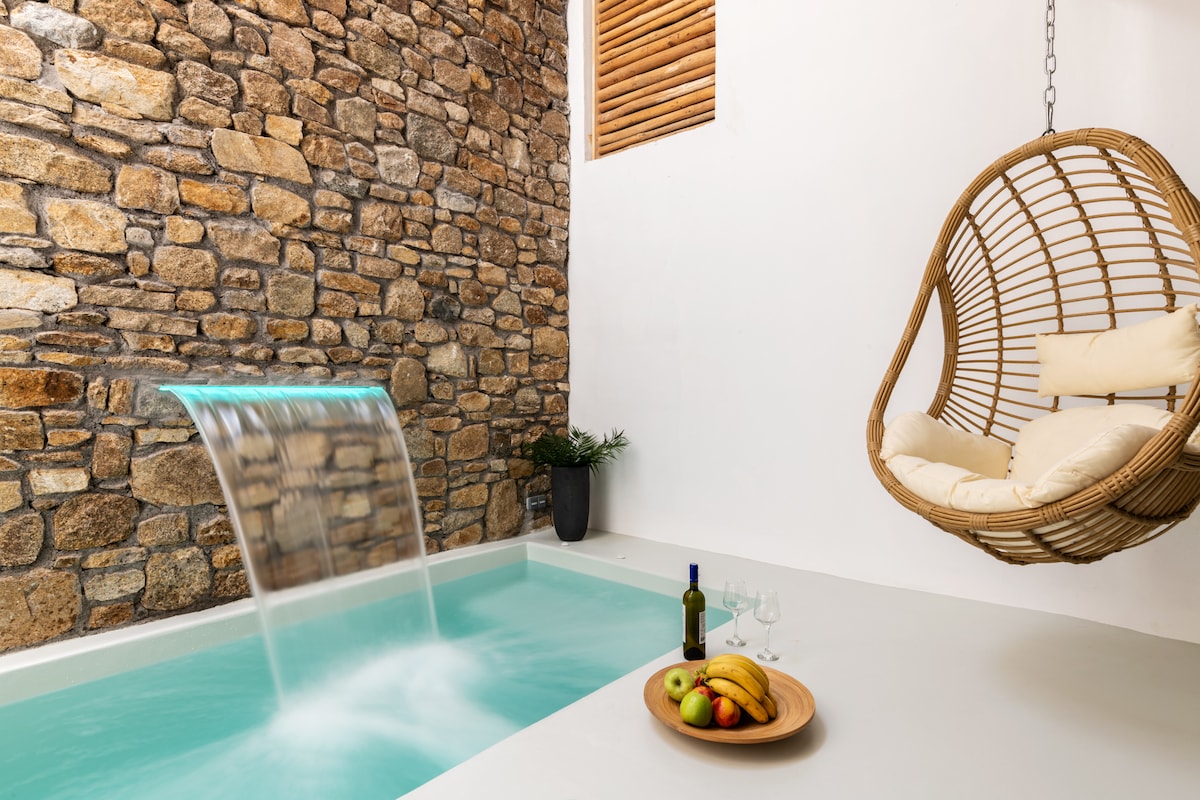 An indoor pool features a gentle waterfall, providing a serene ambiance. A hanging chair with soft cushions complements the space, while a wooden tray holds fresh fruit and a bottle of wine, enhancing the inviting atmosphere of relaxation.