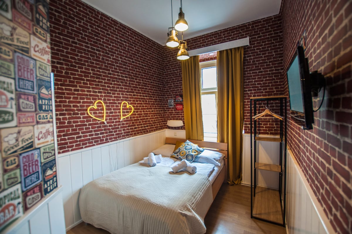 The room features a double bed adorned with decorative pillows, set against a backdrop of exposed brick walls. Warm yellow curtains frame a window, while modern light fixtures hang above. A flat-screen TV is mounted on one wall, and a minimalist shelving unit stands nearby.