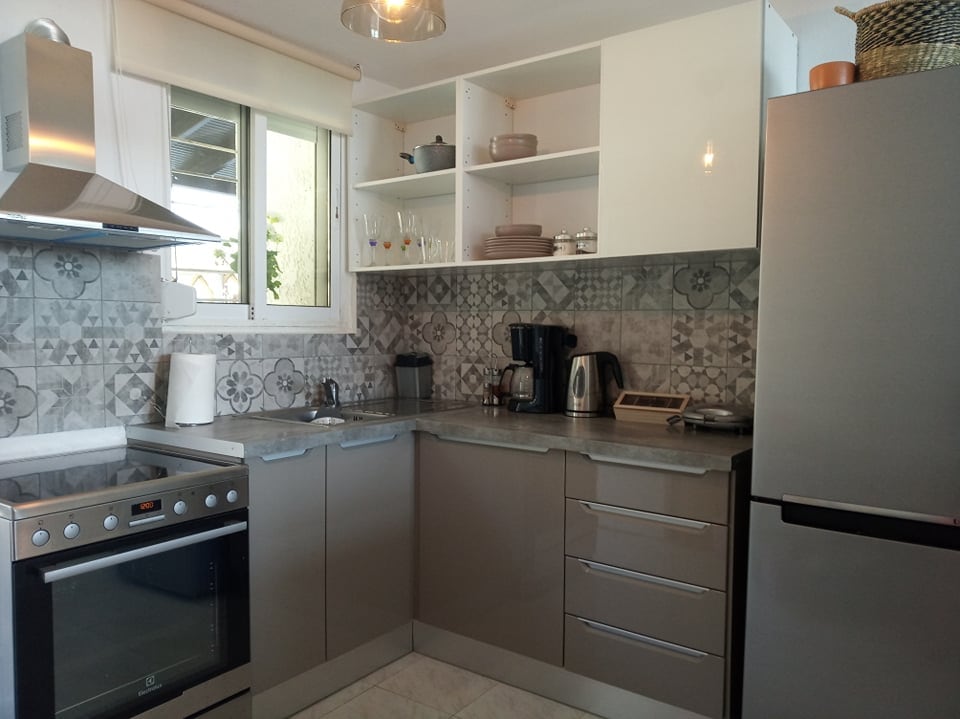 A modern kitchen is equipped with a stainless steel oven, refrigerator, and coffee maker. Neutral-colored cabinets hold dishware and glassware, while a patterned backsplash adds texture. Natural light enters through a window, illuminating the space.