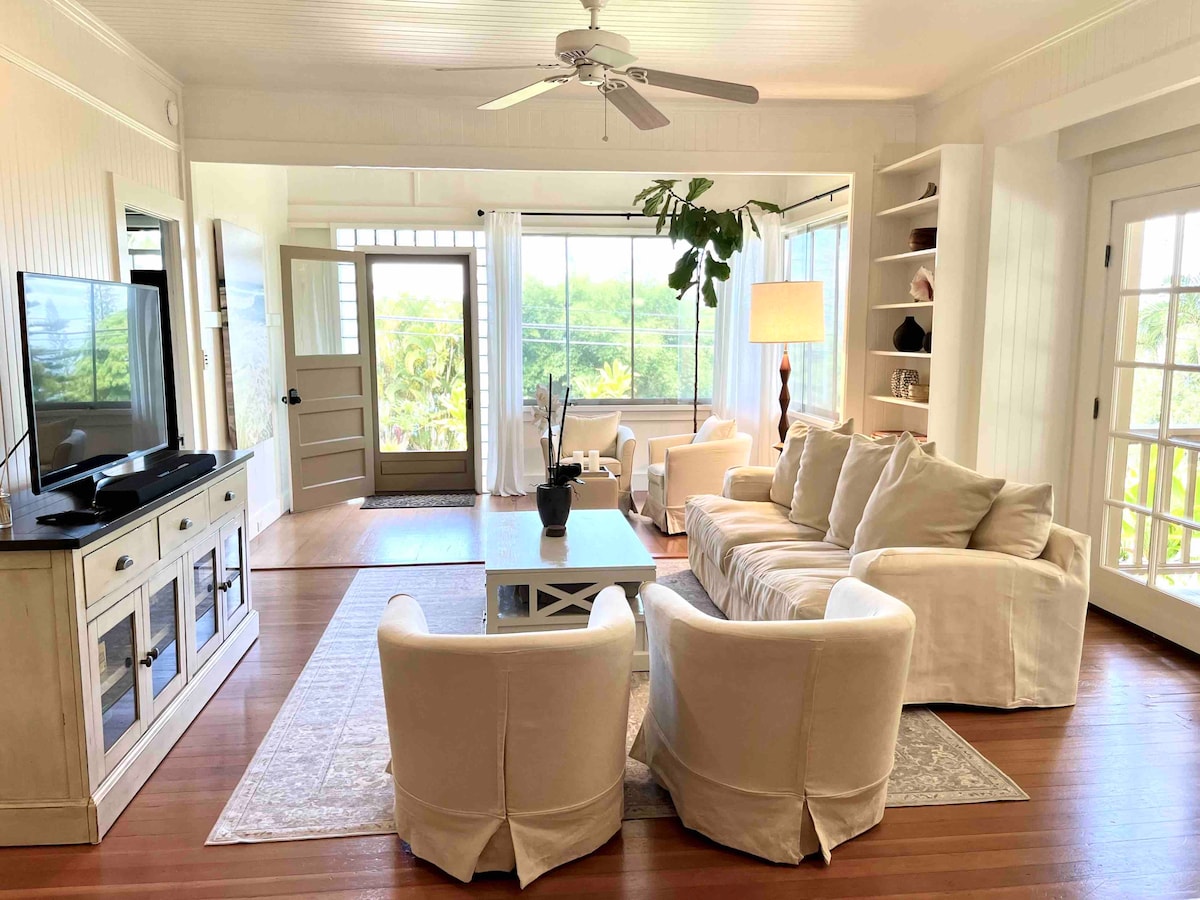 A spacious living area is illuminated by natural light pouring in through large windows. Comfortable seating is arranged around a central coffee table, with a ceiling fan providing a gentle breeze. Soft, neutral decor enhances the calm ambience.