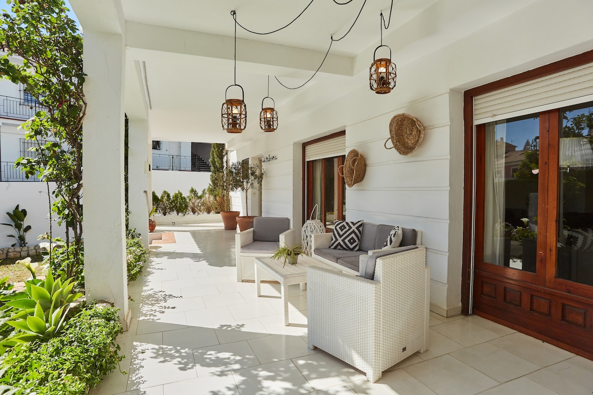 A covered patio area is shown with modern outdoor furniture, including a white wicker sofa and armchairs. Decorative light fixtures hang from the ceiling, while planters with greenery are positioned along the walls, adding a fresh touch to the space.