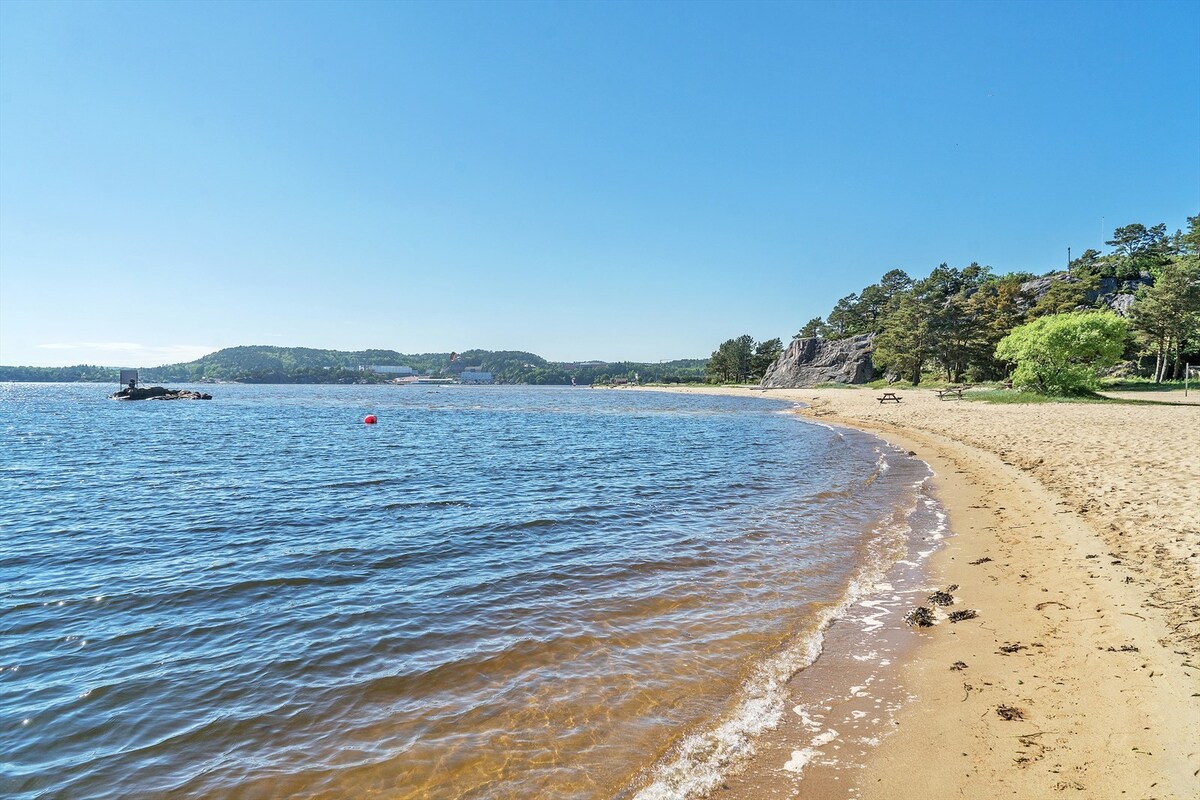 A sandy beach stretches along the shoreline, bordered by calm waters. Gentle waves lap at the shore, while scattered greenery and hills frame the background. The clear blue sky adds to the serene atmosphere, inviting relaxation and outdoor activities.
