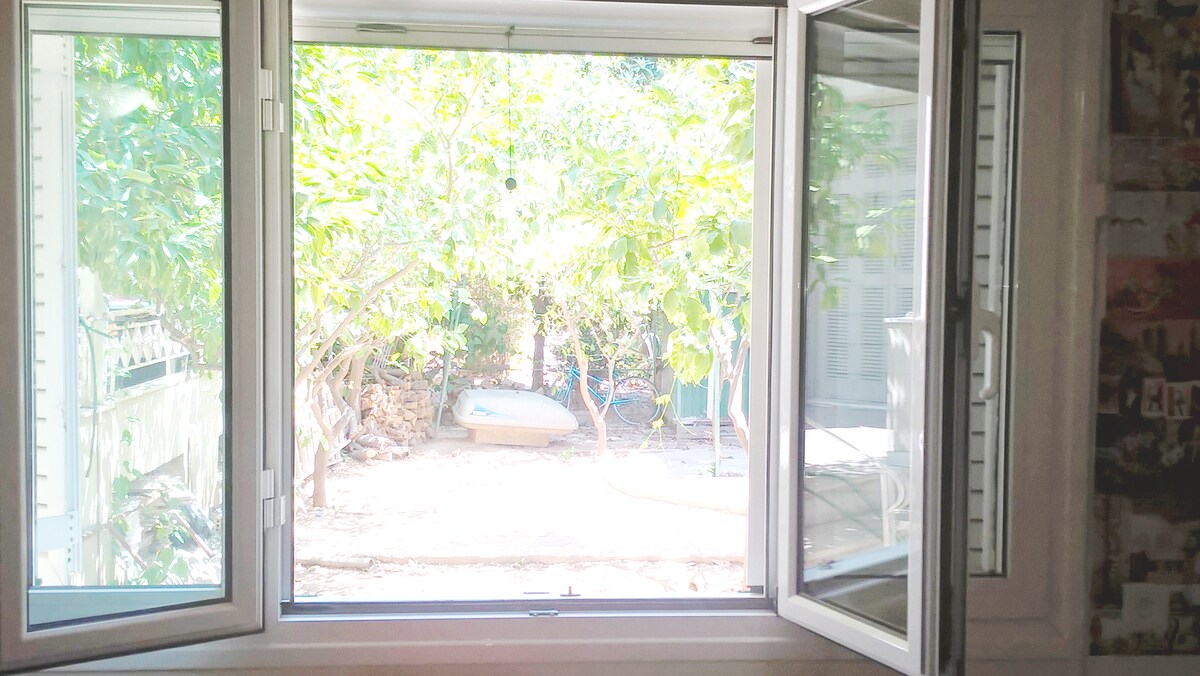 A window is opened to reveal a lush garden filled with greenery. Sunlight filters through the leaves, casting a natural glow on the ground. A serene outdoor space is visible, featuring a pathway and a small boat in the distance.