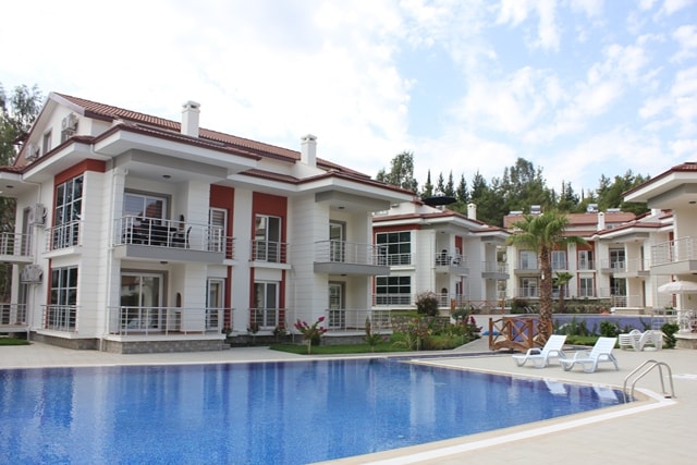 A large swimming pool is showcased in front of two-story apartment buildings, surrounded by well-maintained greenery. Sun loungers are positioned beside the pool, and clear skies are visible above, enhancing the overall inviting ambiance of the outdoor space.