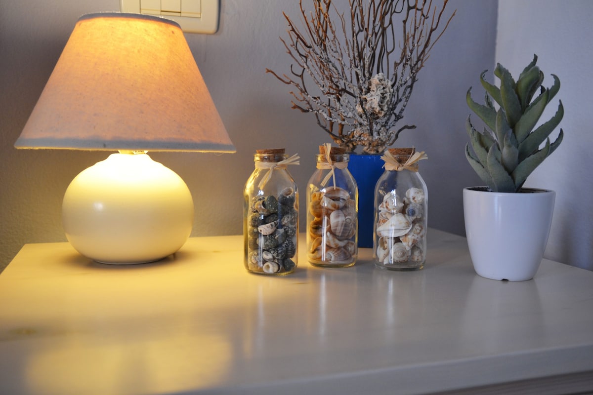 A bedside table is adorned with a soft light from a lamp, casting a warm glow. Three glass jars filled with seashells and stones are arranged alongside a small potted plant and natural branches, adding a touch of coastal decor.