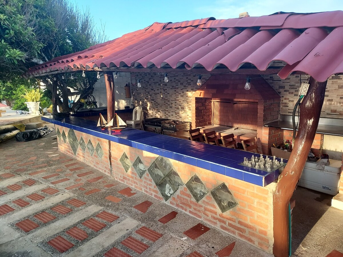 An outdoor cooking area is highlighted by a covered bar with tiled counters and a brick barbecue. Wooden tables and chairs are arranged under the shelter, surrounded by greenery. Soft lighting is suspended from the roof, adding a welcoming touch to the space.