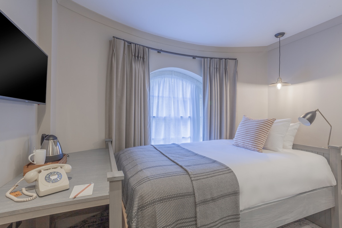 A warm sleeping area features a spacious bed with white linens and a patterned throw blanket. Natural light filters through an arched window, draped with light curtains. A small table holds a kettle and an assortment of amenities, while a flat-screen TV is mounted on the wall.