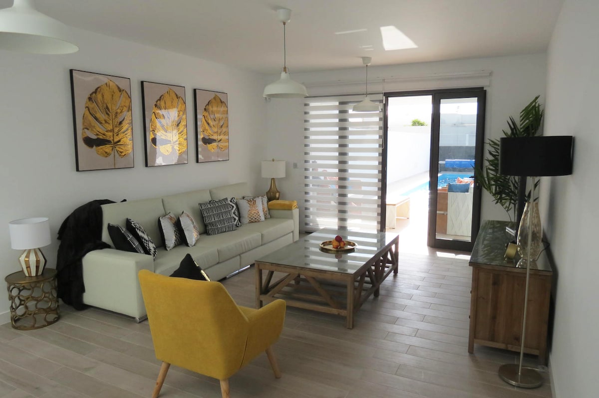 The spacious living area features a light color palette with a large sectional sofa adorned with various cushions. A coffee table sits atop wooden flooring, accompanied by a decorative lamp. Double doors lead to the outdoor terrace, enhancing natural light within the space.
