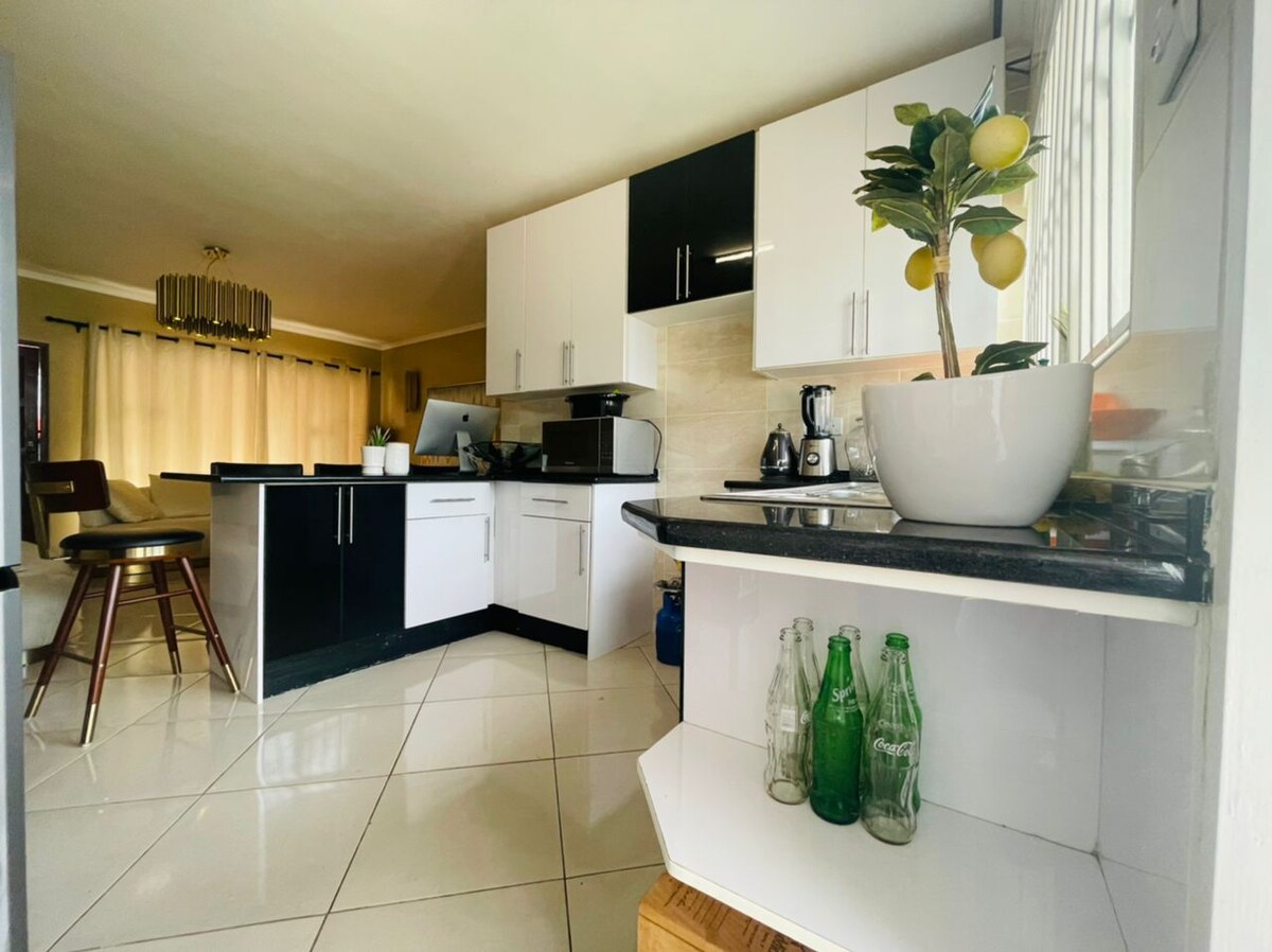 A modern kitchen is presented with sleek black and white cabinetry, featuring a spacious countertop and various appliances. Natural light enters through the large window, illuminating decorative plants and glassware on the shelves, creating a clean and functional cooking space.