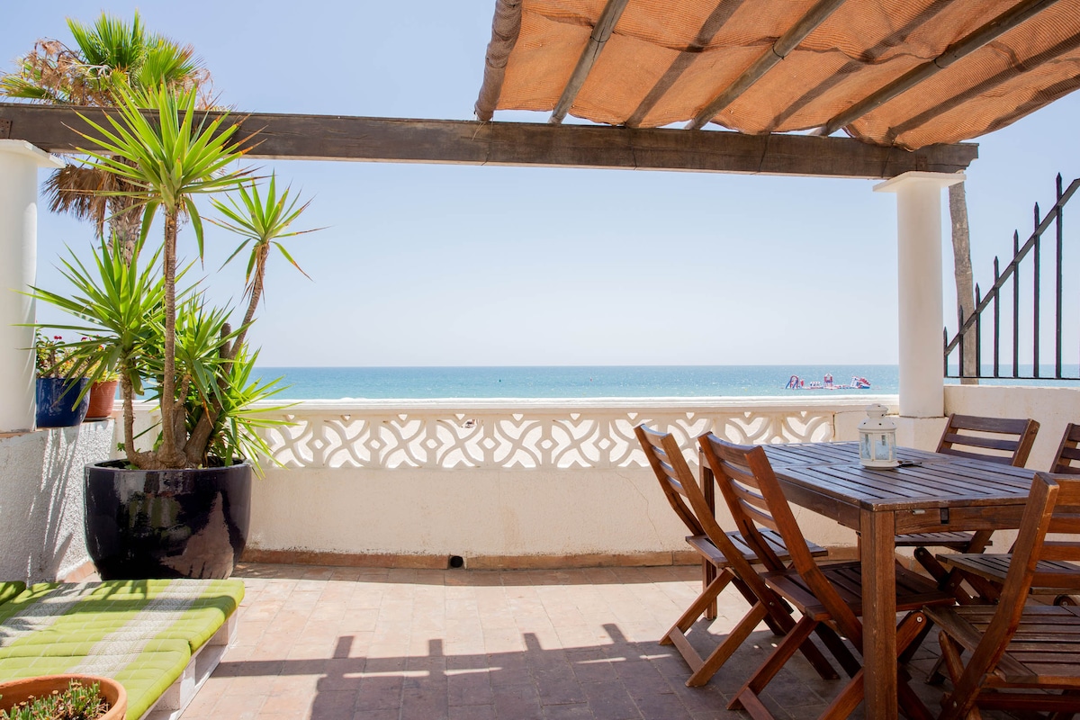 A terrace with a direct view of the sea features outdoor furniture, including a wooden dining table with matching chairs. Potted plants are placed along the edge, and a pergola provides shade. The calm waters and clear sky are visible in the background.