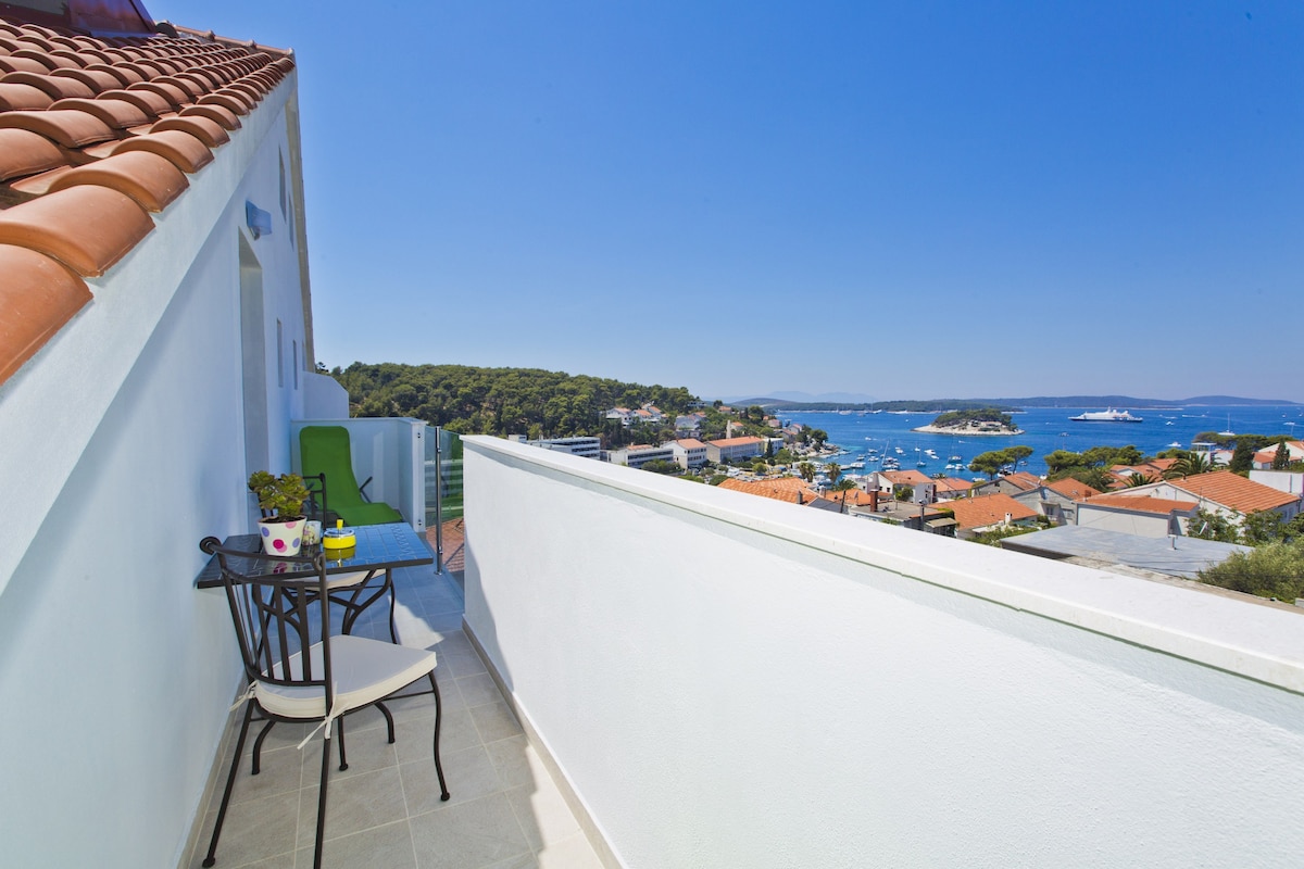 A private balcony showcases a stunning sea view overlooking the Pakleni islands. Two chairs and a small table are positioned on the tiled surface, providing a serene spot to enjoy the surroundings. The blue sky creates a bright backdrop against the greenery and coastal scenery.
