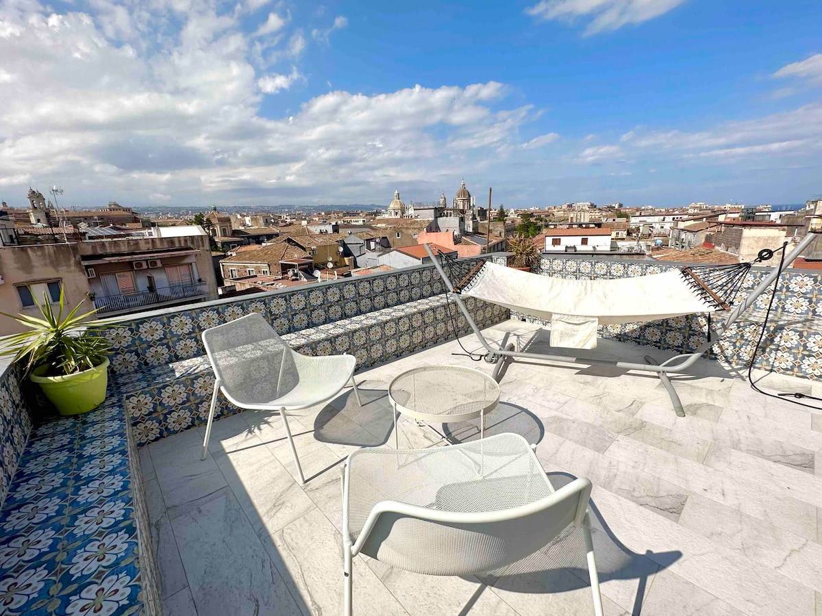 A private terrace features sleek seating options, including two chairs and a table, alongside a comfortable hammock. Decorative tiles adorn the perimeter, providing a colorful touch. The expansive view showcases rooftops and notable landmarks of Catania beneath a partly cloudy sky.