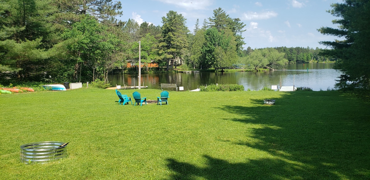A spacious green lawn borders the serene waterfront, featuring five blue adirondack chairs arranged around a fire pit. Kayaks and paddle boats are visible along the shore, set against a backdrop of lush trees and calm water reflecting the sky.