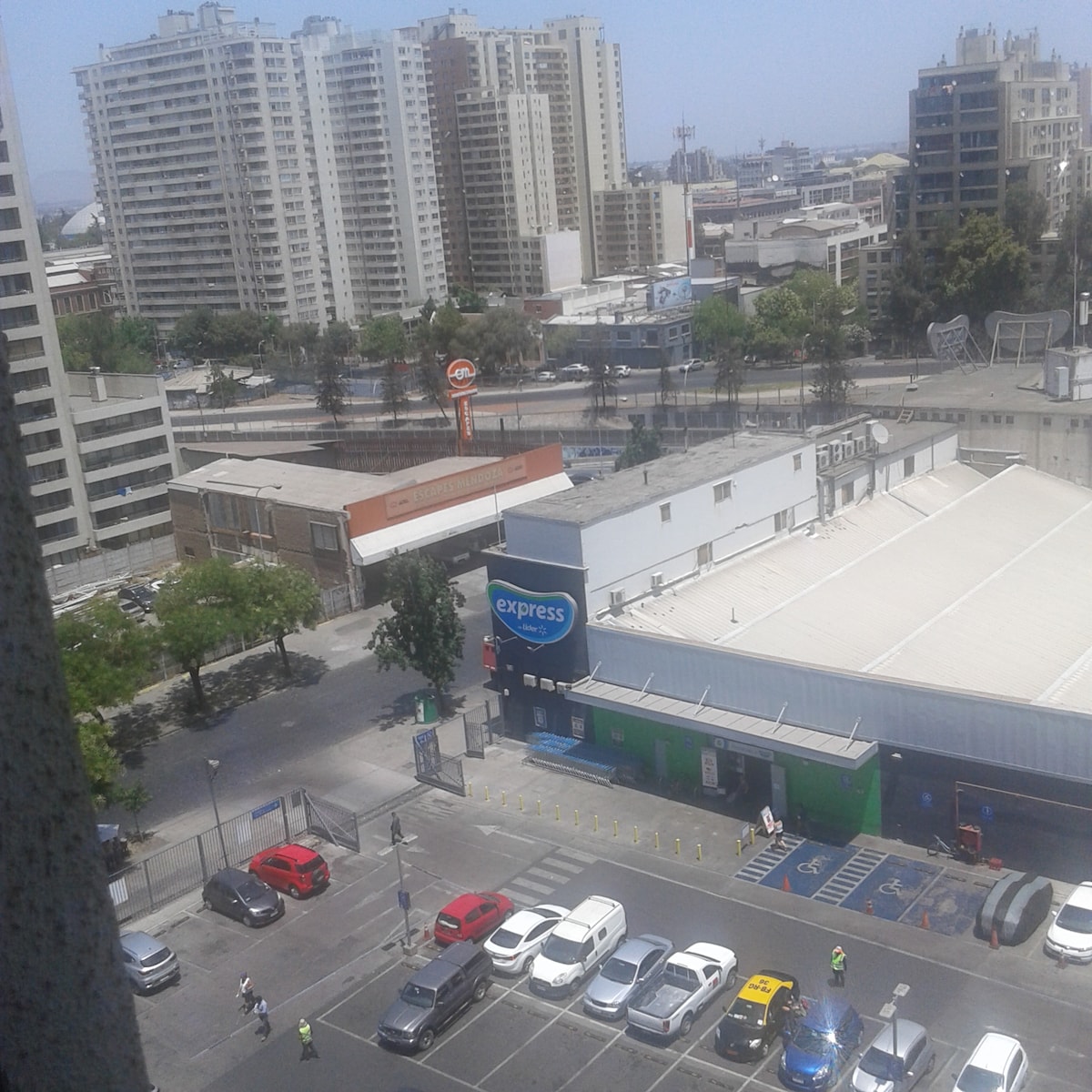 A panoramic view captures the surrounding cityscape, featuring tall buildings and a bustling street below. An express store is visible, along with a parking area filled with vehicles. The vibrant urban environment is punctuated by trees lining the street.