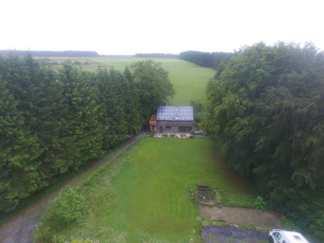 The image presents an aerial view of a chalet surrounded by lush greenery, with solar panels on the roof. A spacious lawn extends towards the horizon, framed by dense trees. A path leads to the property, highlighting the peaceful rural setting.