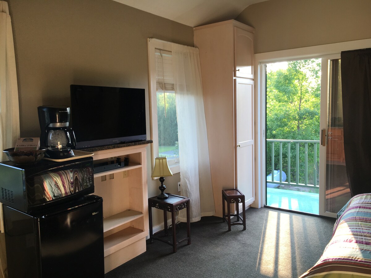 A bright queen room features a kitchenette area with a mini refrigerator and a coffee maker on a wooden console. Sunlight illuminates the space through large windows, highlighting the two small seating stools beside the bed. A glass door provides access to a small deck overlooking greenery.