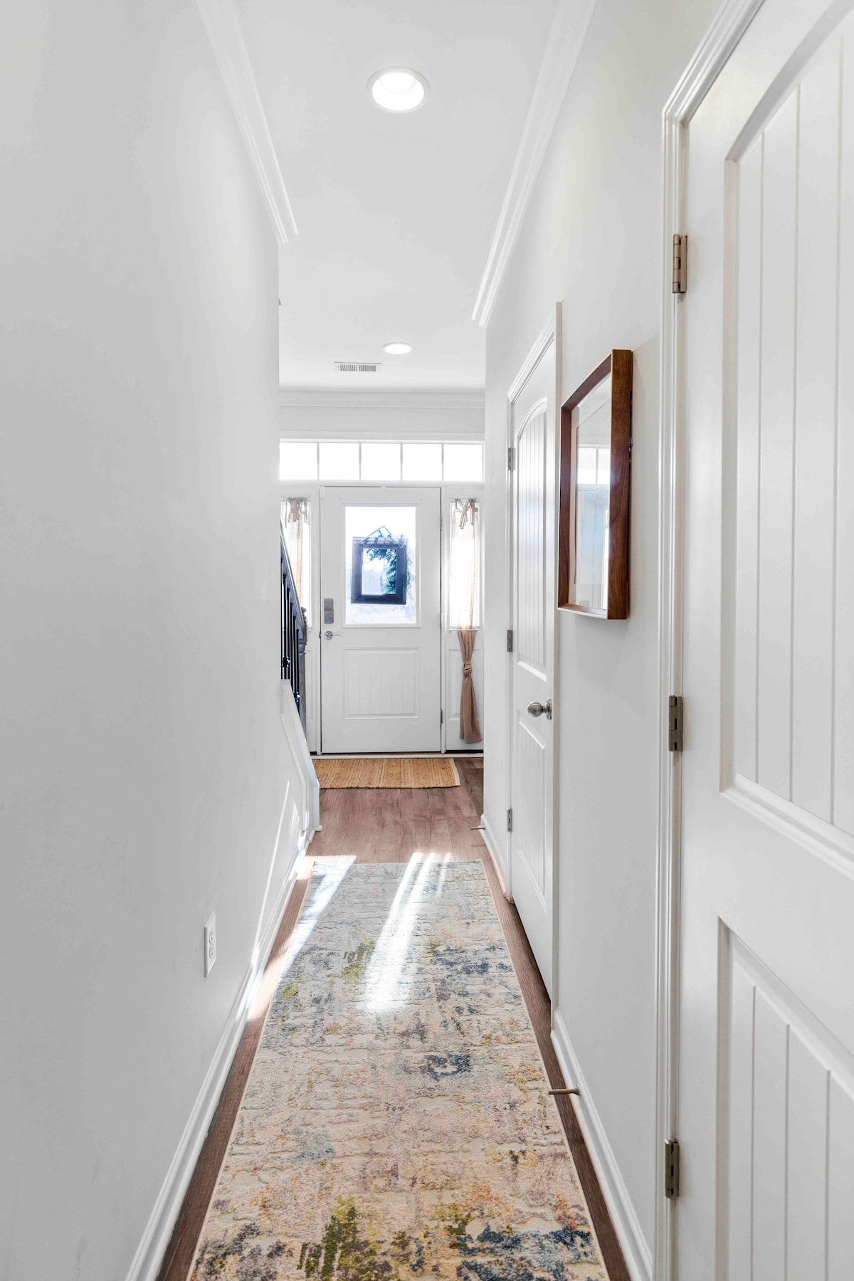 A bright hallway features smooth, light-colored walls and a warm-toned wooden floor. A decorative runner rug adds color, while natural light filters through a frosted glass door at the end. A small framed artwork is mounted on the wall to the right.