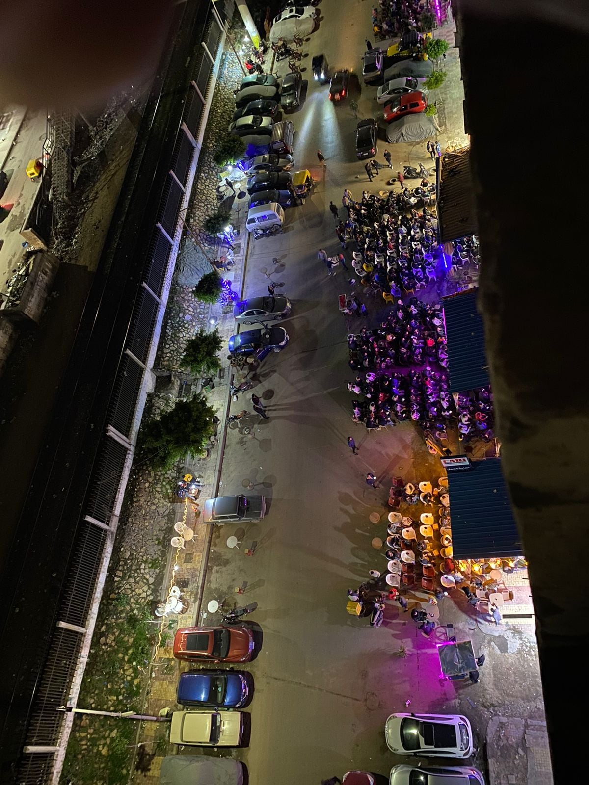 An aerial view captures a bustling street scene at night, illuminated by colorful lights. Rows of parked cars line the sides, while tables filled with patrons are seen below. The surrounding area features greenery along the edges, contributing to the lively atmosphere.