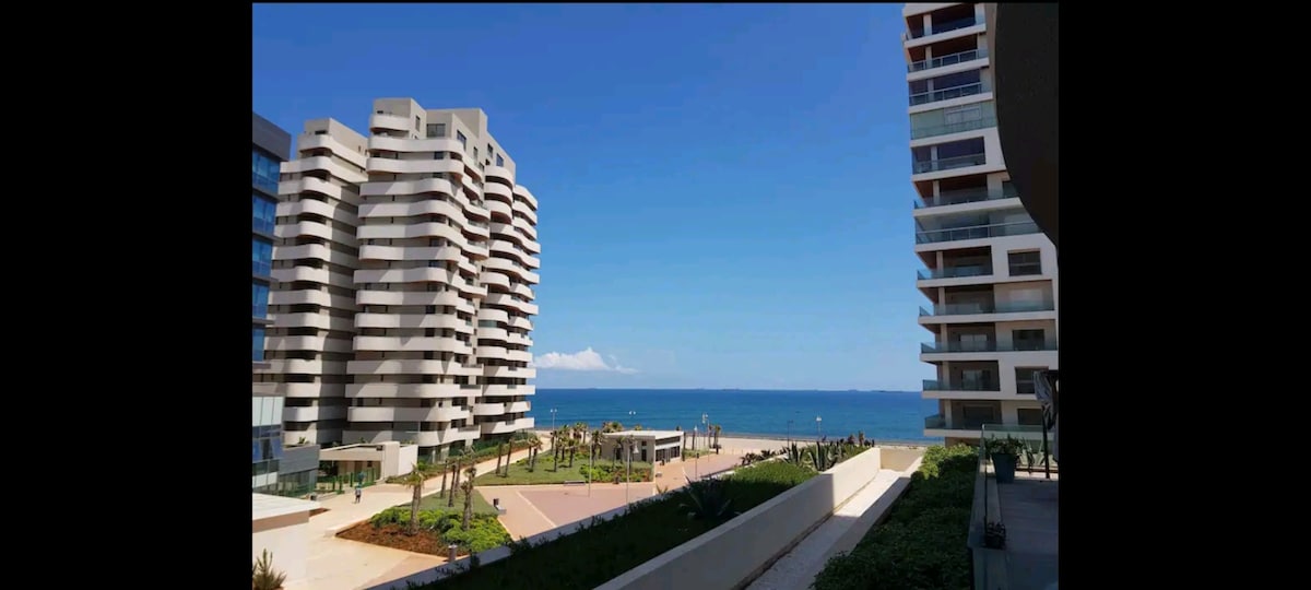A clear view of the ocean is framed by modern buildings. The shoreline is lined with palm trees, and a spacious area for leisure activities is visible in the foreground. A bright blue sky extends above the scene, creating an open and airy atmosphere.