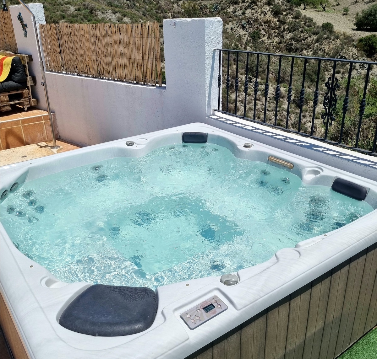 A relaxing jacuzzi is situated on a wooden platform, with crystal clear water bubbling gently. The surrounding area features a low fence and a scenic view of the mountainous landscape, creating a serene atmosphere for unwinding and enjoying the natural surroundings.
