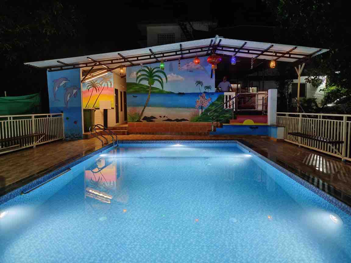 An illuminated swimming pool reflects a vibrant mural of a beach scene, featuring palm trees and waves. Colorful lanterns hang above a covered area, which includes seating. The inviting blue water is complemented by surrounding light tiles, providing a relaxing ambiance.
