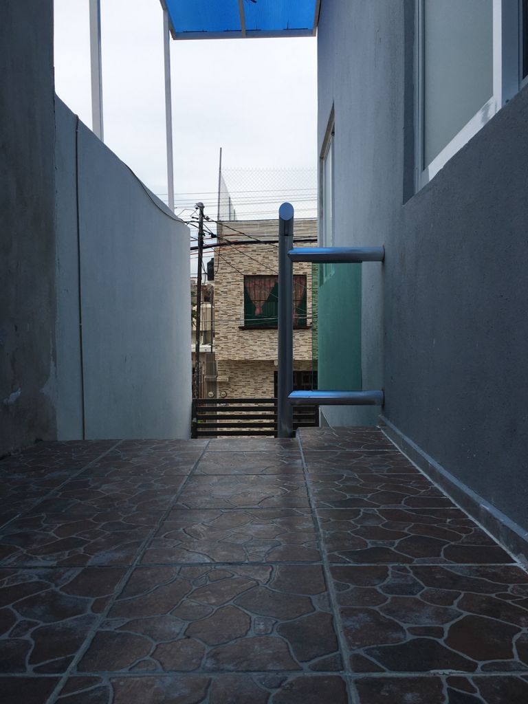 A narrow corridor is visible, featuring a textured floor with rectangular tiles. Light gray walls frame the space, leading toward a blue sky above. In the distance, the neighboring building is partially seen through a window, providing a view of an urban setting.