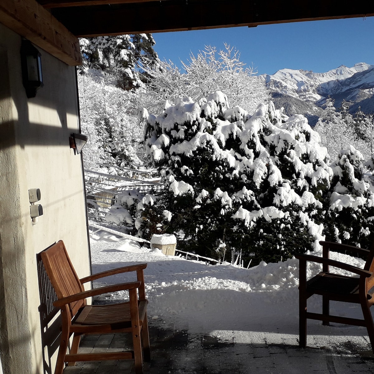 A private terrace is shown, featuring two wooden chairs positioned for mountain views. Snow-covered trees and a winter landscape fill the background, while sunlight brightens the scene against the blue sky.