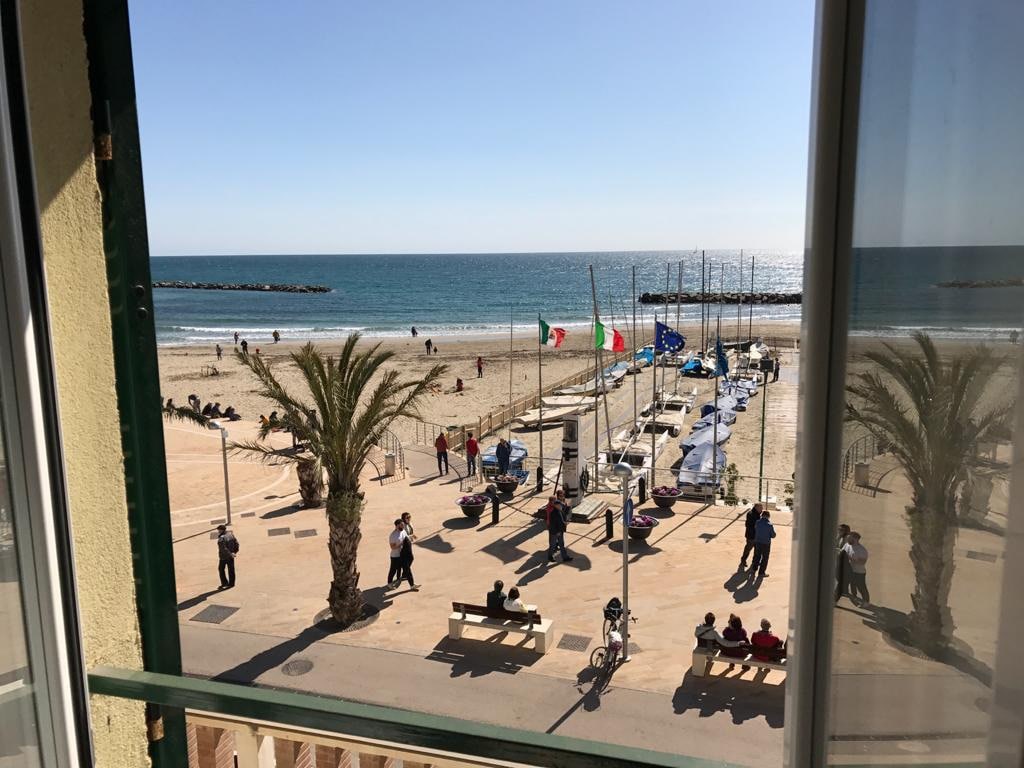 A view of the beach is visible from the window, showcasing golden sand and clear blue waters. Numerous people are strolling along the promenade, while flags flutter near the beach. Palm trees line the walkway, adding a tropical feel to the coastal scene.
