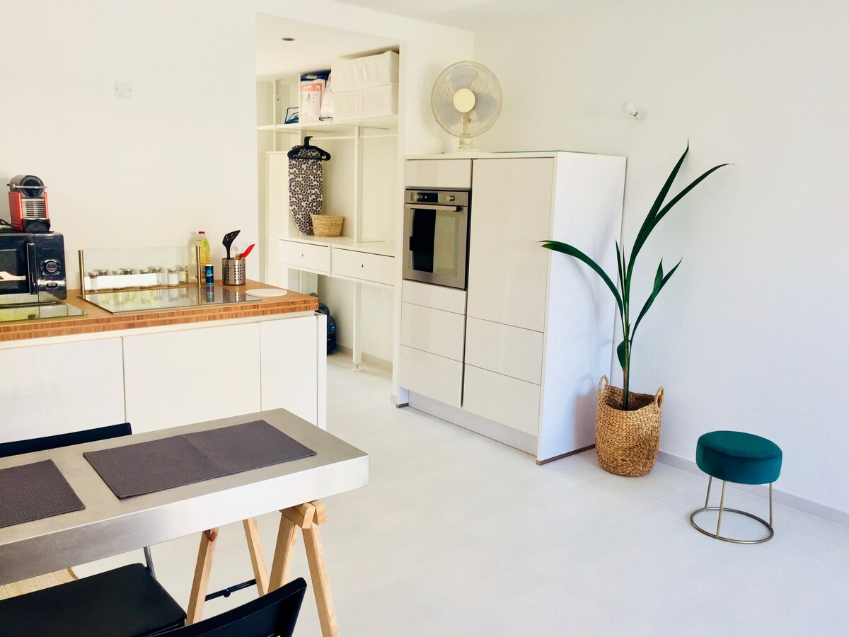 A bright kitchen area features modern cabinetry and appliances, complemented by a small dining table and chairs. A tall houseplant adds a touch of greenery, while light-colored tiles enhance the spacious feel of the room. The overall design emphasizes cleanliness and functionality.