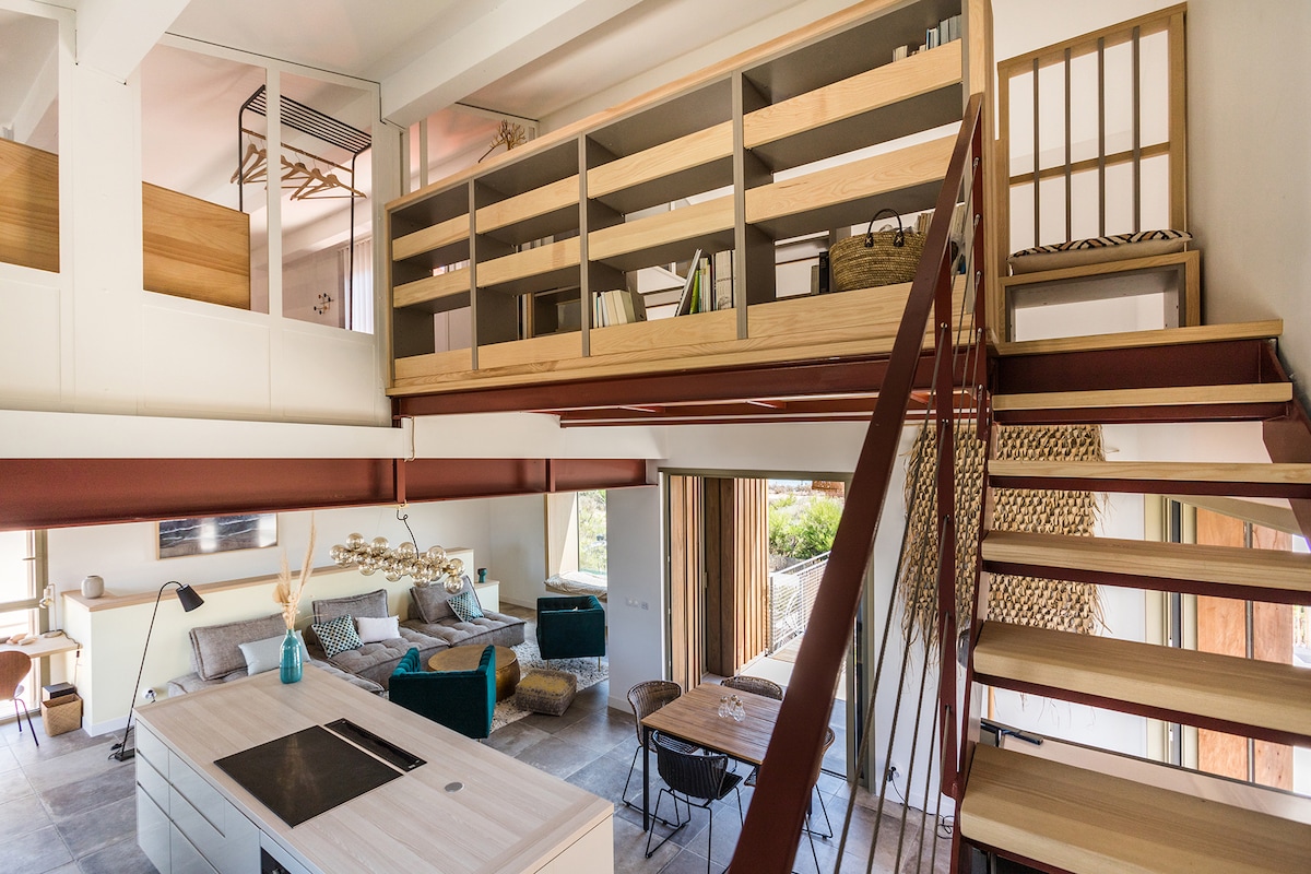 An airy interior space showcases a wooden staircase leading to an upper loft area. The open-plan living area features a modern kitchen island and a cozy seating arrangement. Natural light floods in through large windows, highlighting the minimalist decor and built-in shelving.