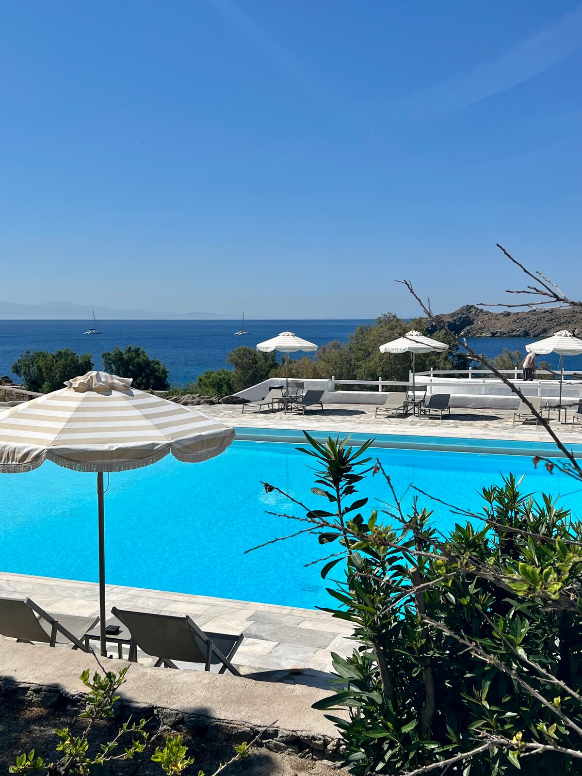 A serene outdoor swimming pool is framed by lush greenery, showcasing clear turquoise water. Umbrellas provide shade over lounge chairs positioned around the pool, with calm ocean views in the background, and distant hills set against a clear blue sky.