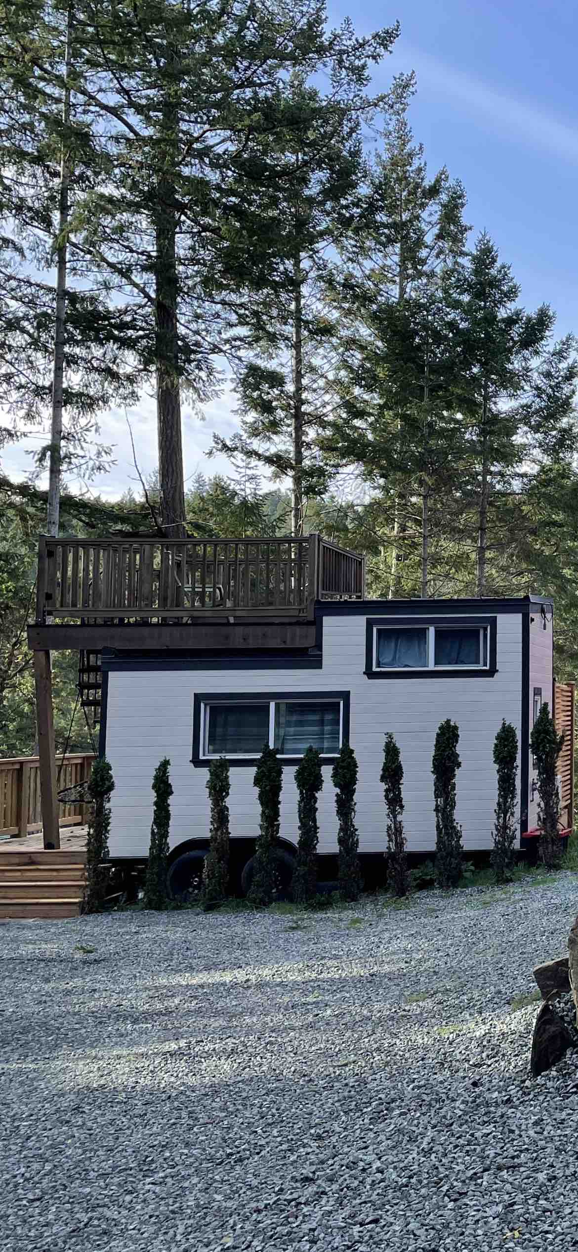 A tiny house with a modern exterior is showcased, featuring large windows and a raised deck. The structure is situated among tall trees, with a pathway flanked by neatly trimmed shrubs leading to the entrance. A gravel space surrounds the house.